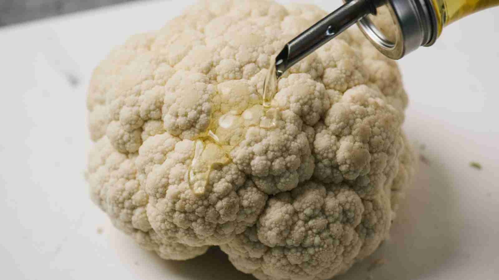 A bottle pours olive oil onto a whole cauliflower placed on a white surface.