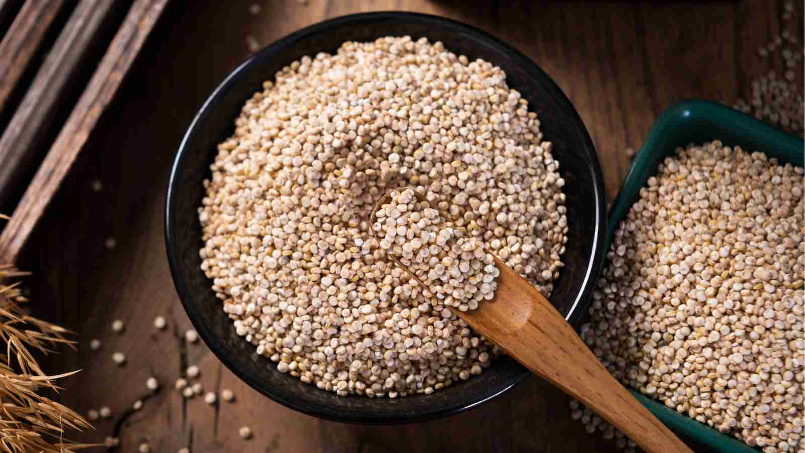 A wooden spoon rests in a black bowl filled with quinoa grains on a wooden surface.
