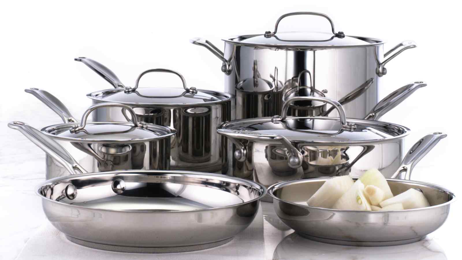 A set of stainless steel cookware including pots, pans, and a few pieces of chopped onion in one pan, placed on a white surface.