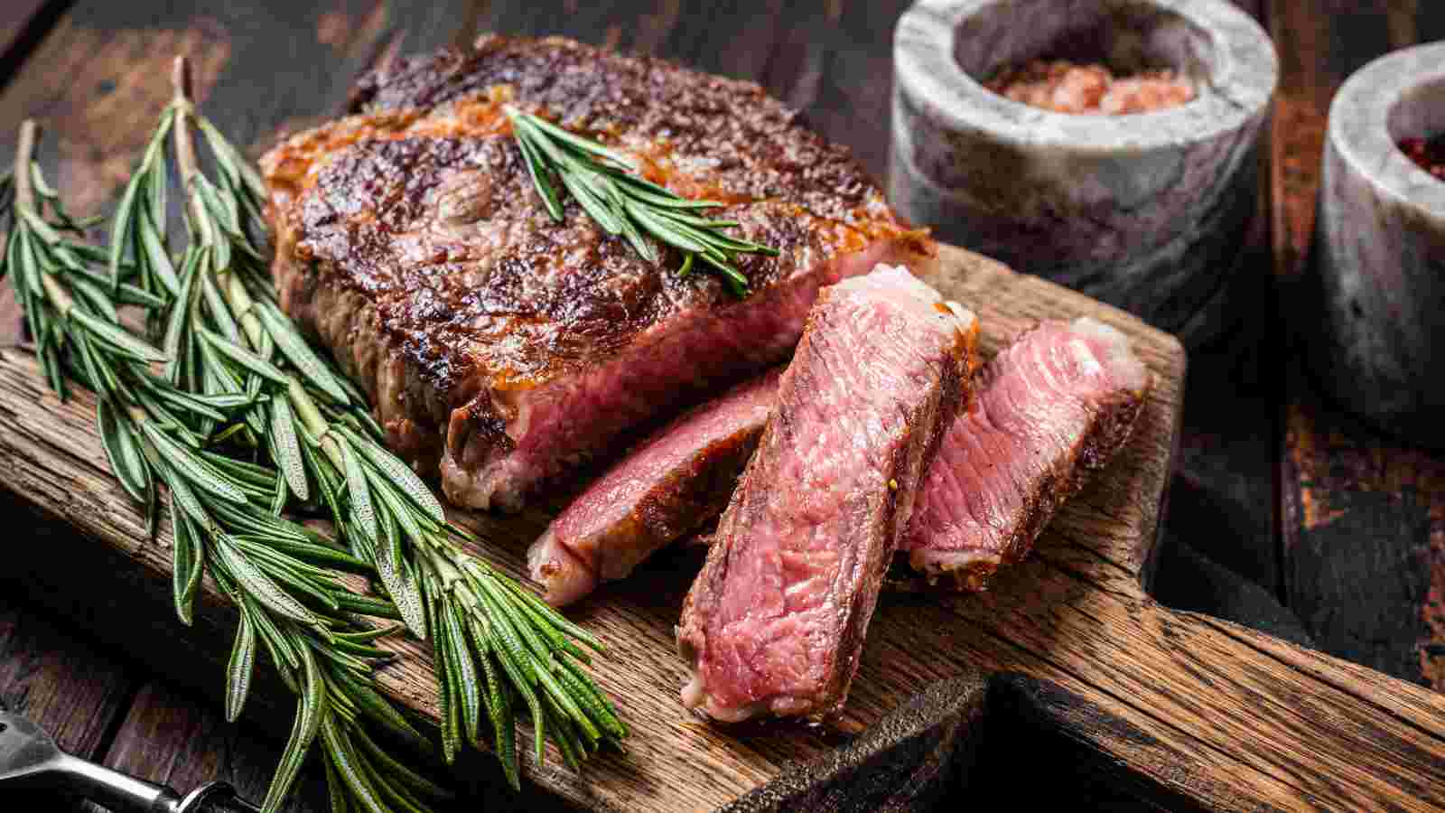 A cooked steak, partially sliced, garnished with rosemary sprigs, sits on a wooden cutting board.