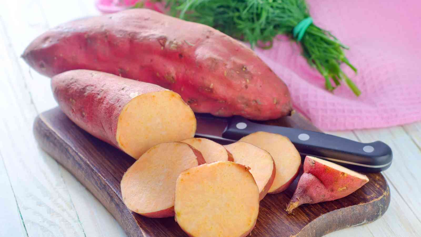 Sliced sweet potatoes and a knife on a cutting board with a larger, whole sweet potato and a bunch of green leaves in the background.