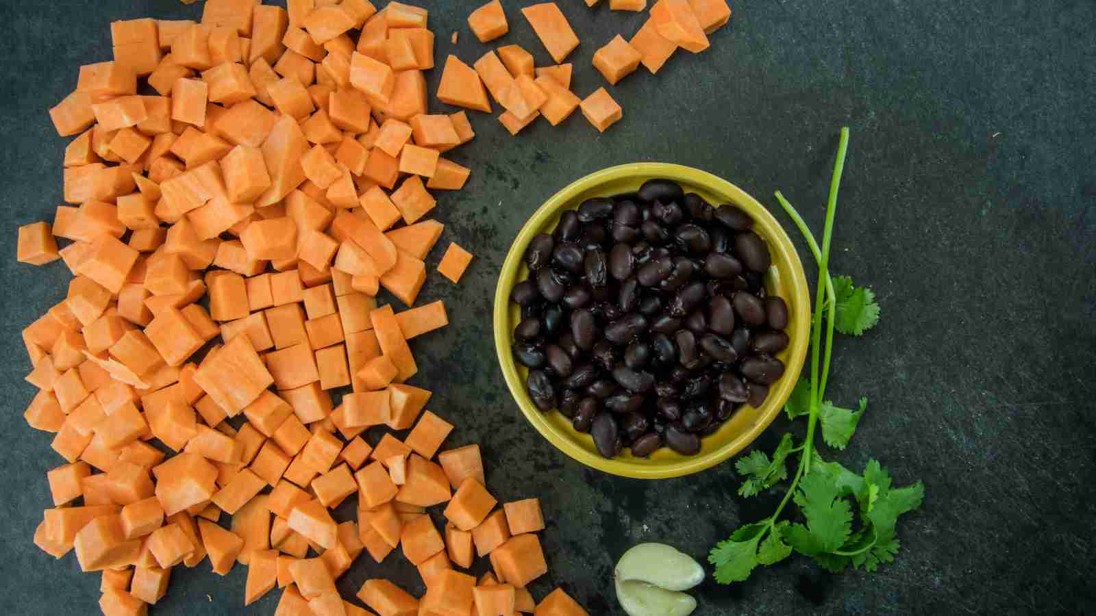 Chopped sweet potatoes on a dark surface beside a bowl of black beans, with cilantro and garlic cloves.
