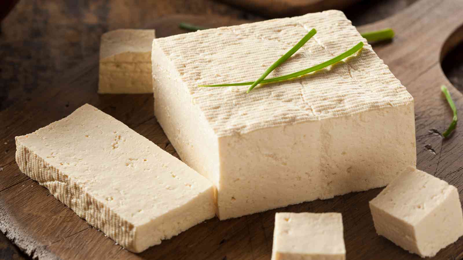 A block of firm tofu placed on a wooden surface, garnished with a few green herbs.