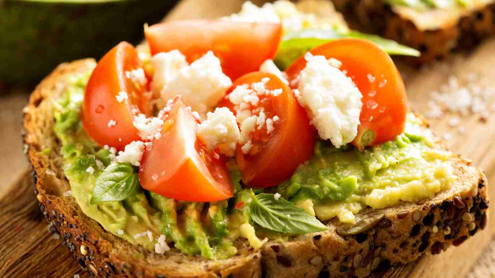 A slice of multigrain toast topped with mashed avocado, halved cherry tomatoes, crumbled cheese, and fresh basil leaves.