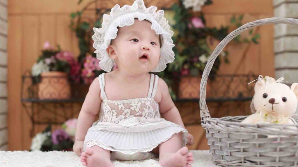 A baby wearing a lace bonnet and dress sits next to a wicker basket with a stuffed bear.