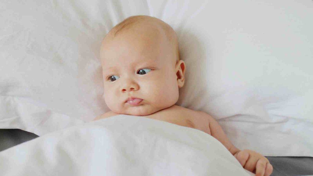 A baby with a neutral expression lies on a white bed, partially covered by a white blanket.