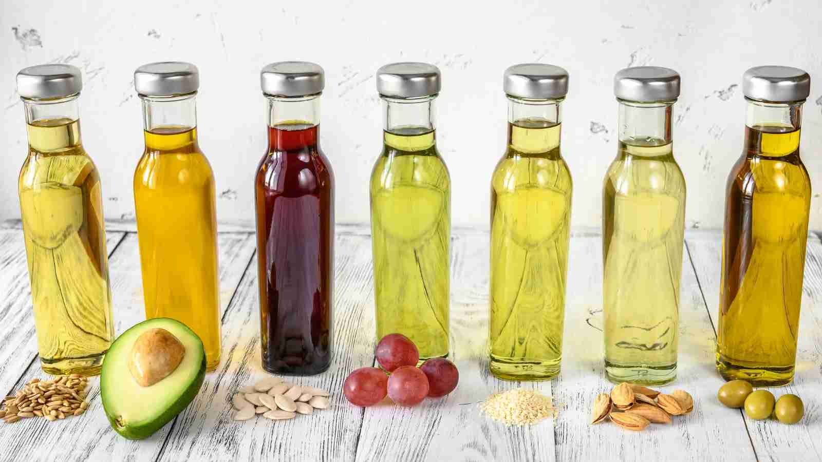 Seven bottles of various cooking oils are lined up with ingredients like avocado, grapes, seeds, nuts, and olives in front of them on a white wooden surface.