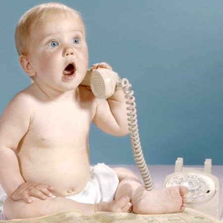 A baby with blonde hair, wearing only a diaper, is sitting on a pink surface and holding a beige rotary phone's receiver to its ear, looking surprised. An open book is in front of the baby.