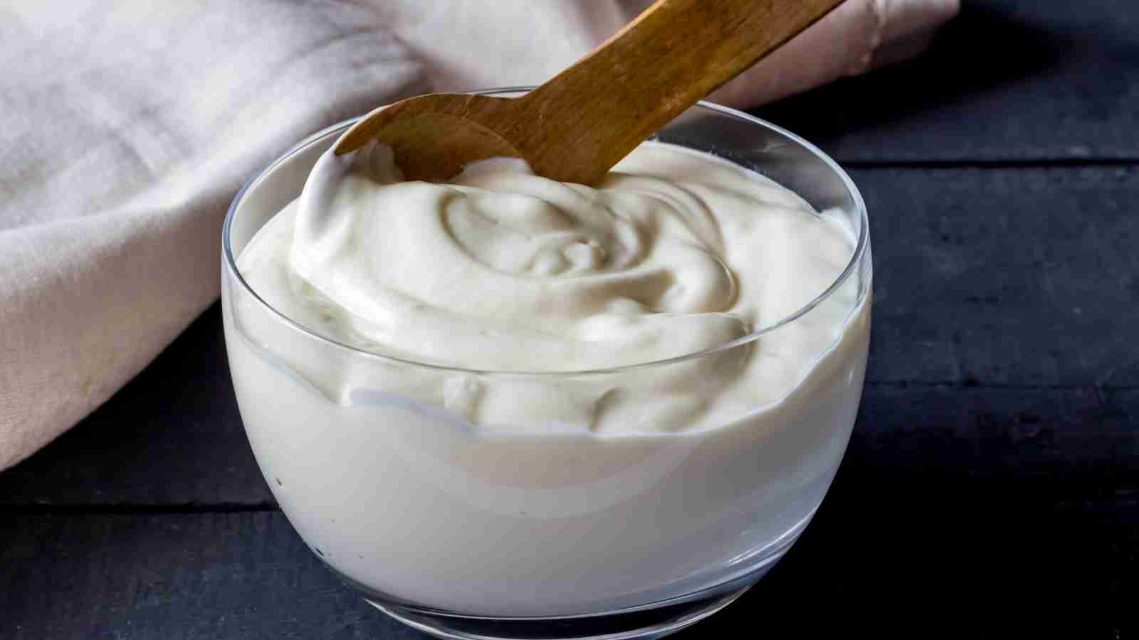 A glass bowl filled with creamy yogurt and a wooden spoon resting inside it. There is a cloth napkin in the background.