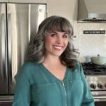 A woman with shoulder-length gray hair and bangs smiles in a kitchen, standing in front of a refrigerator and stove.