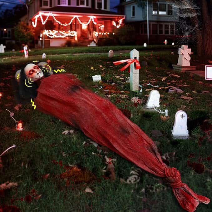 A Halloween display shows a person in a red sack on a lawn surrounded by gravestones, with a house decorated with orange lights in the background.