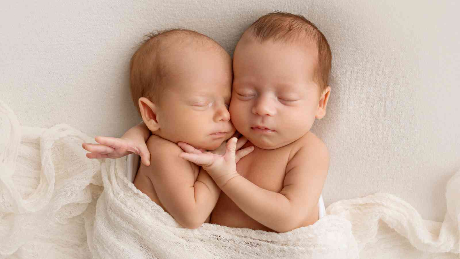 Two newborn babies are lying side by side on a soft white blanket. They are wrapped in a light fabric and have their eyes closed, appearing to sleep peacefully with their heads touching.