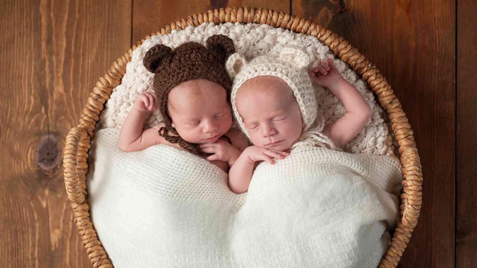Two sleeping babies are cuddled up in a round wicker basket, each wearing a knitted hat with bear ears.