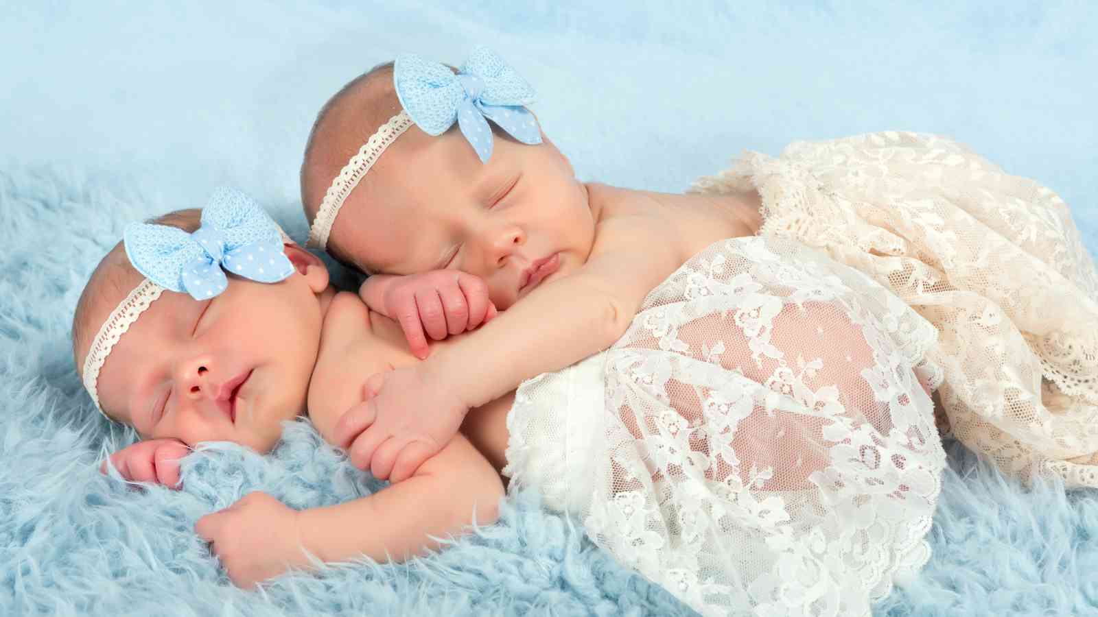 Twin baby girls laying on a blue fua fur sheet wearing laced skirt and matching headbands.