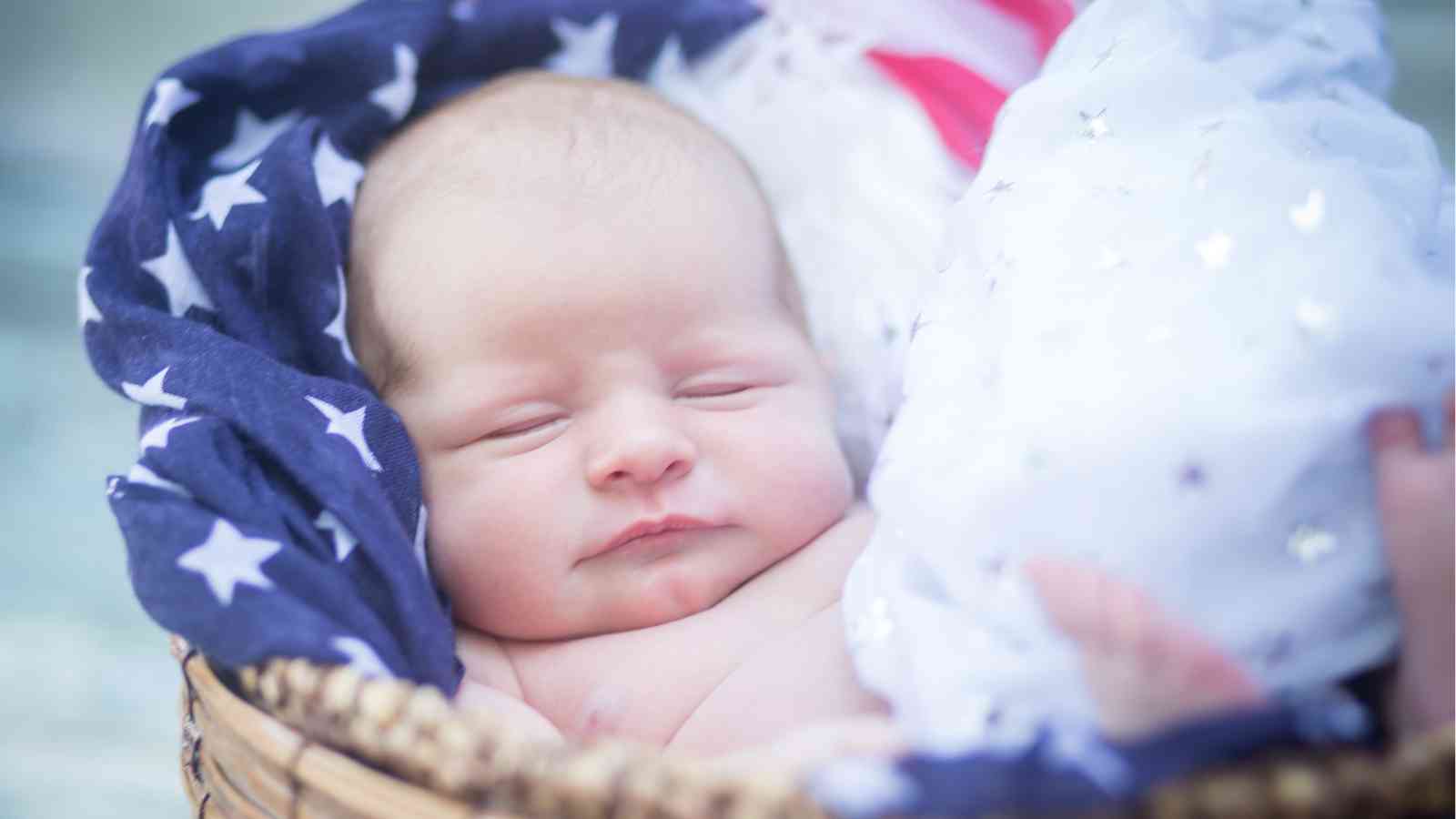 A newborn baby laying in a basket with a US flag underneath his body.