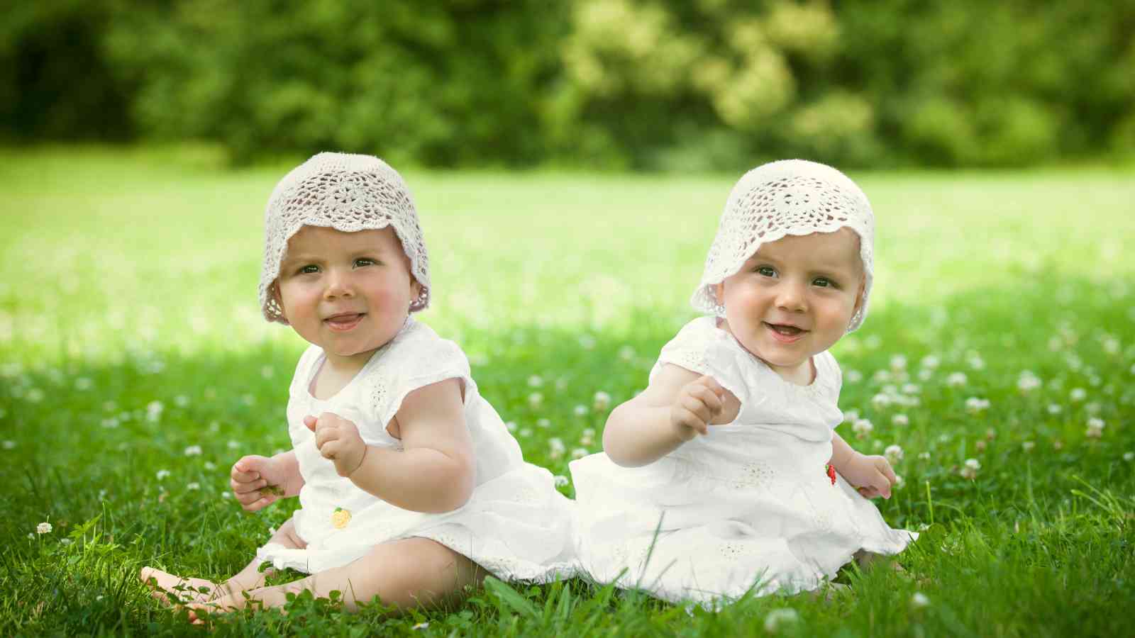 Twin baby girls sitting on the grass wearing matching white dress and hat.