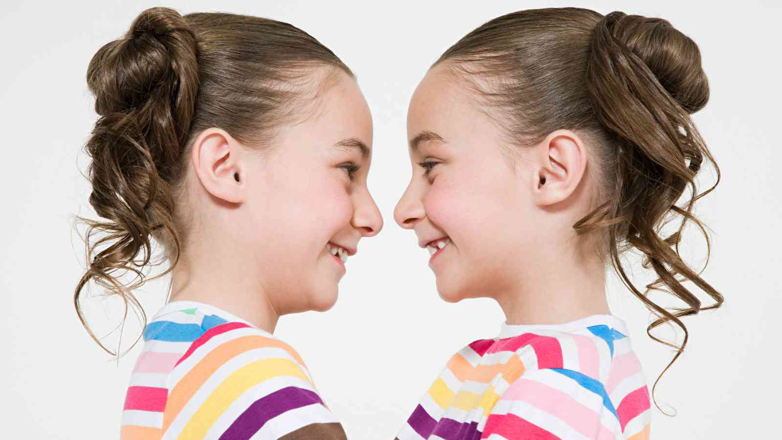 Two young girls with curly hair, wearing rainbow striped shirts, are facing each other and smiling.