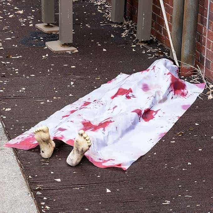 Image of a body covered with a blood-stained sheet, feet exposed, laying on the ground next to a brick wall and wooden structures.