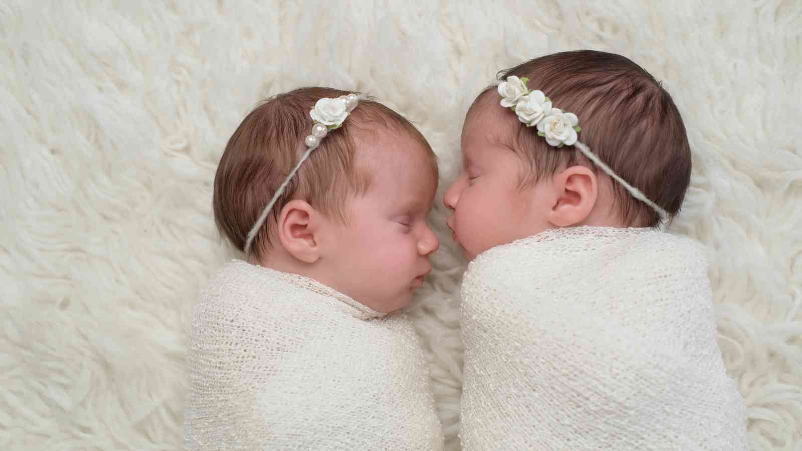 Two newborn baby girls are sleeping soundly, side by side, on a white fuzzy blanket. They are wearing white flower headbands and are wrapped in white blankets.