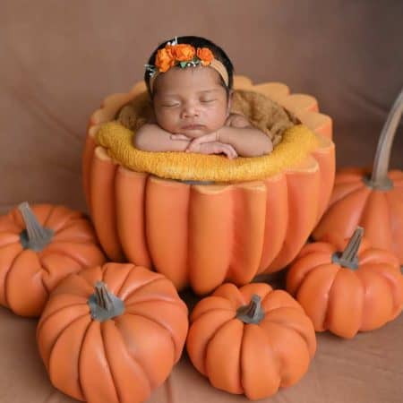 A baby sleeping inside a large, hollow pumpkin prop, with smaller pumpkin props surrounding. The baby wears a floral headband and leans on the edge of the large pumpkin.