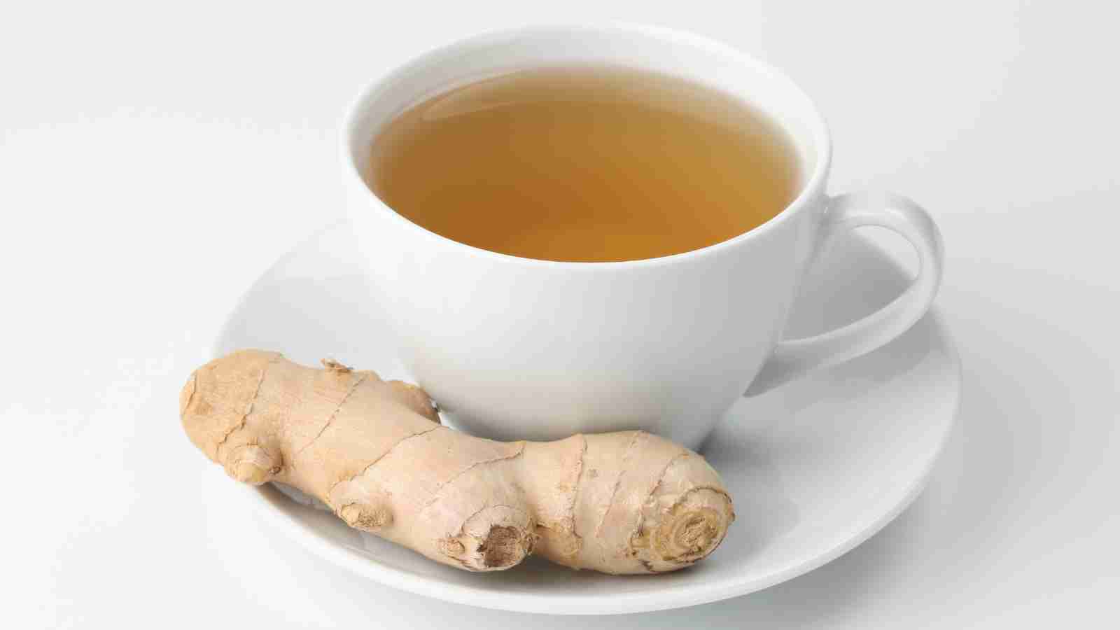 A white cup filled with ginger tea sits on a matching saucer with a piece of fresh ginger root placed next to it.