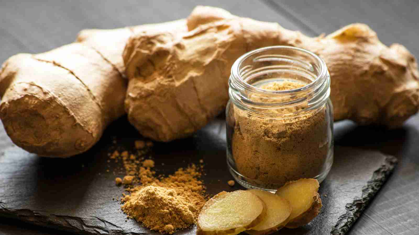Whole ginger root, ground ginger, and sliced ginger arranged on a black slate surface