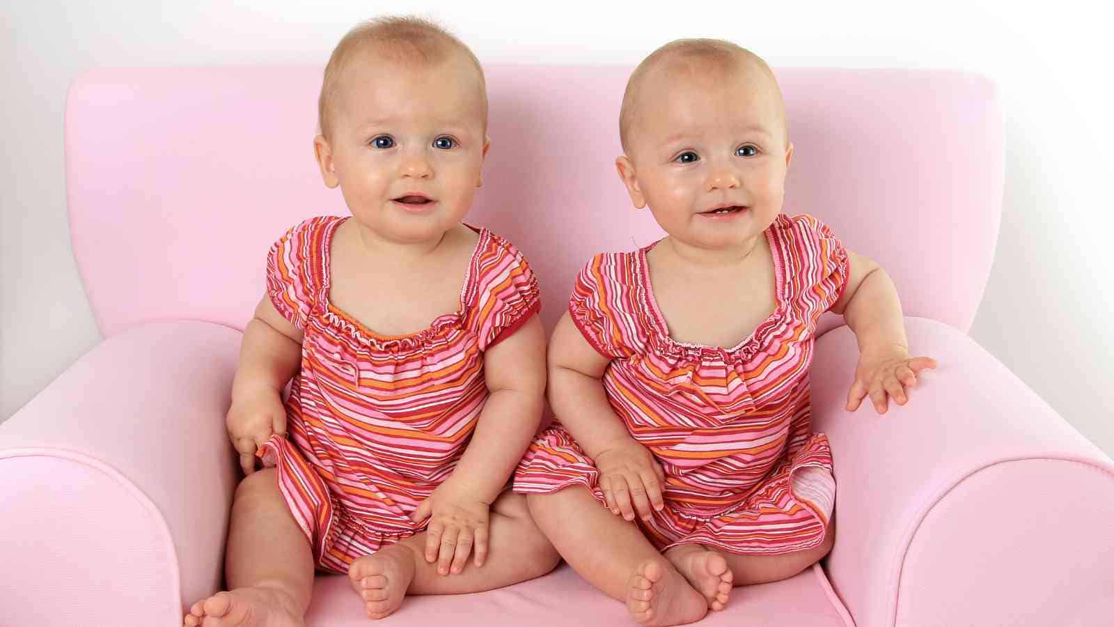 Two baby girls, wearing matching pink and orange striped dresses, are sitting on a pink armchair.