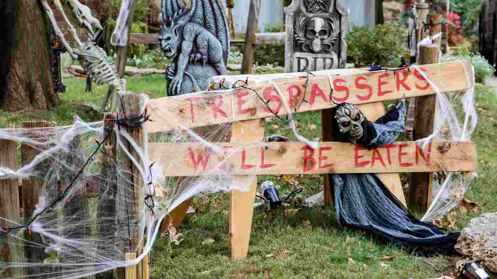 A Halloween display with a wooden sign reading "TRESPASSERS WILL BE EATEN," cobweb decorations, a gargoyle, tombstones, and a hooded skeleton figure.