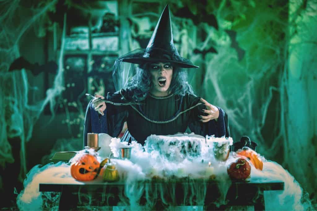 A person dressed as a witch with a pointed hat and green face paint poses behind a table decorated with Halloween items.