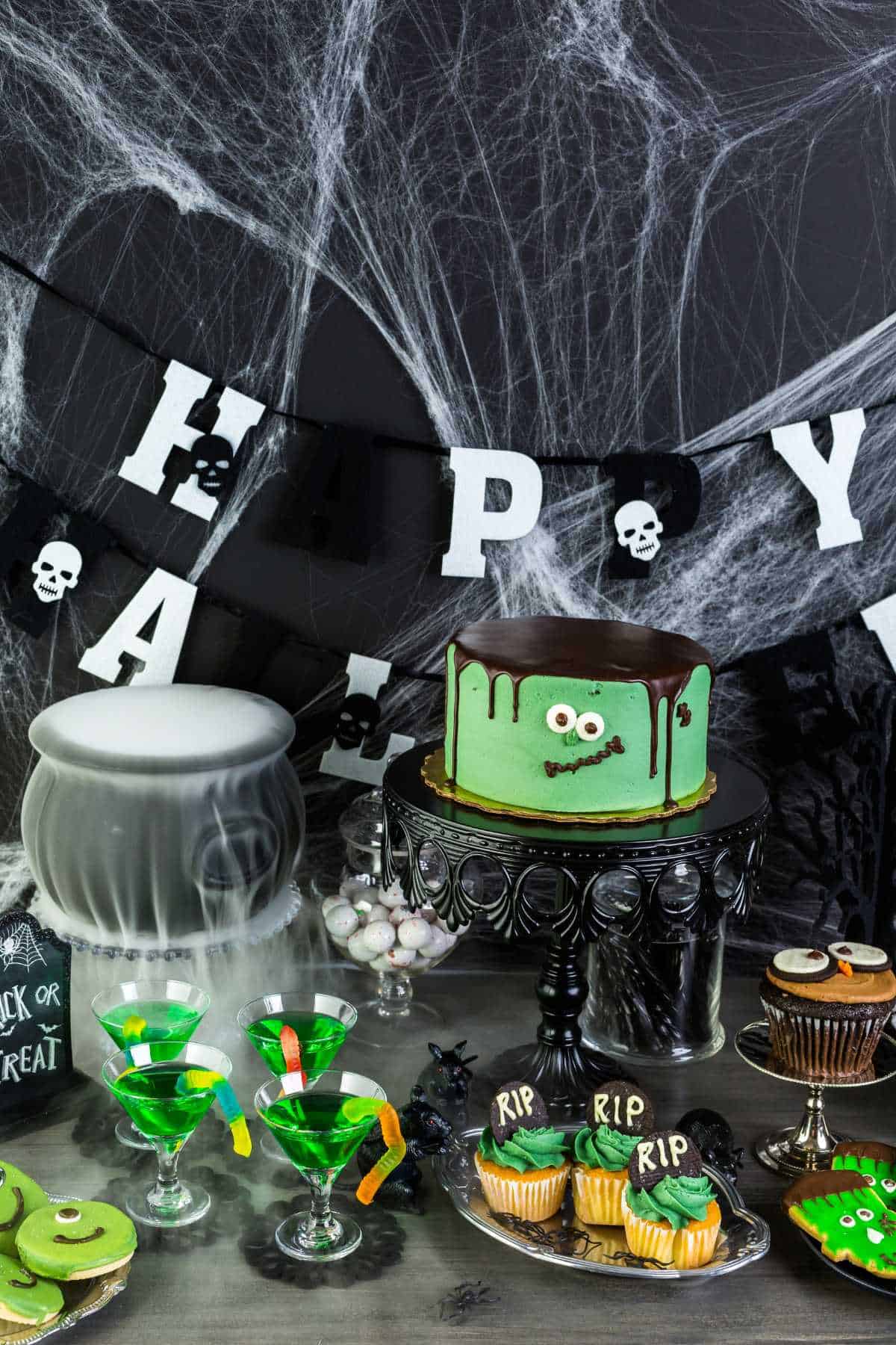 A Halloween-themed table displays a green cake with eyes, spooky cupcakes and cookies, green cocktails, and eerie decorations like skulls, ghostly mist, and a "Happy Halloween" banner.