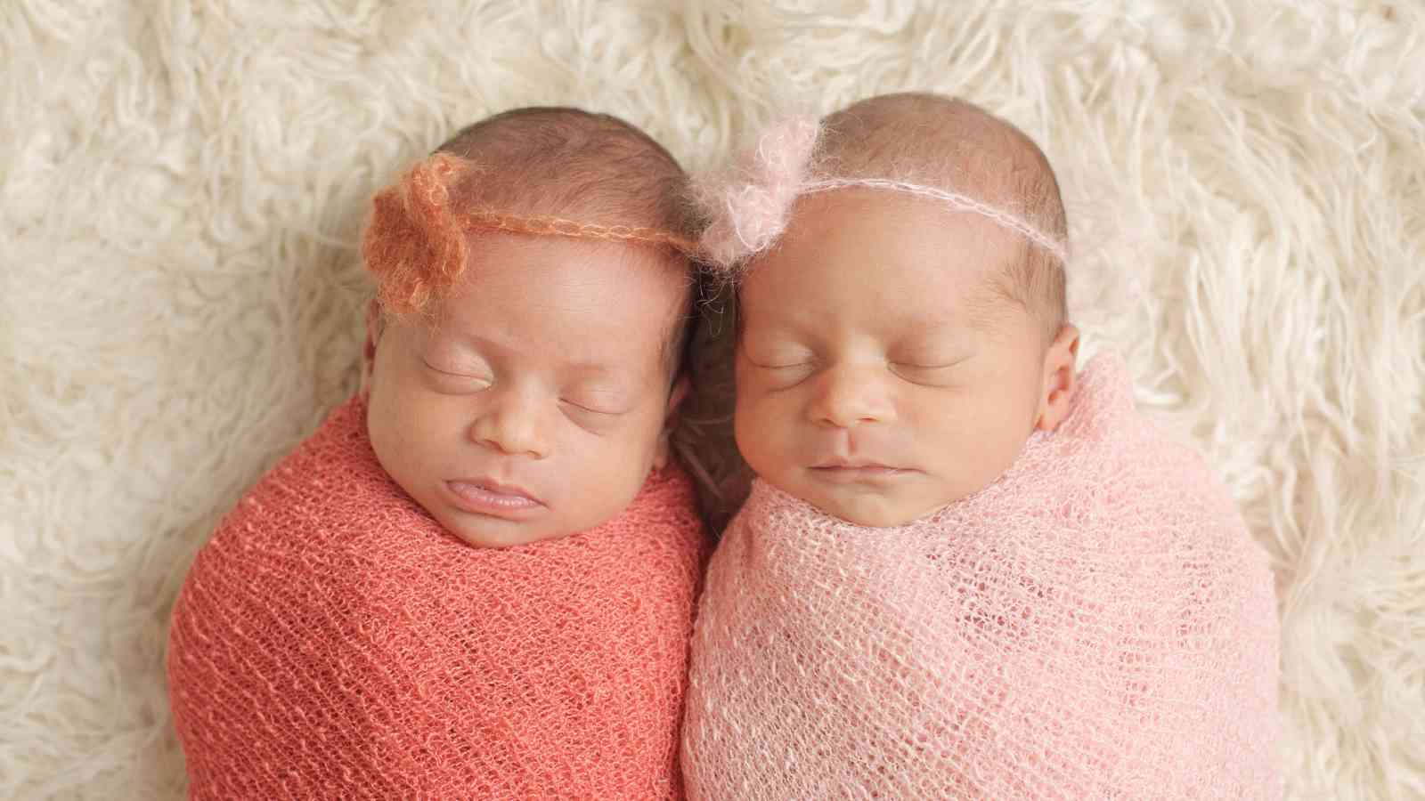 Two newborn baby girls are sleeping soundly, side by side, on a white fuzzy blanket. They are wrapped in blue and green swaddling blankets and are wearing flower headbands.