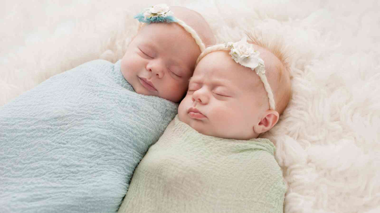Two newborn baby girls are sleeping soundly, side by side, on a light blue blanket. They are wearing matching white outfits and flower headbands.