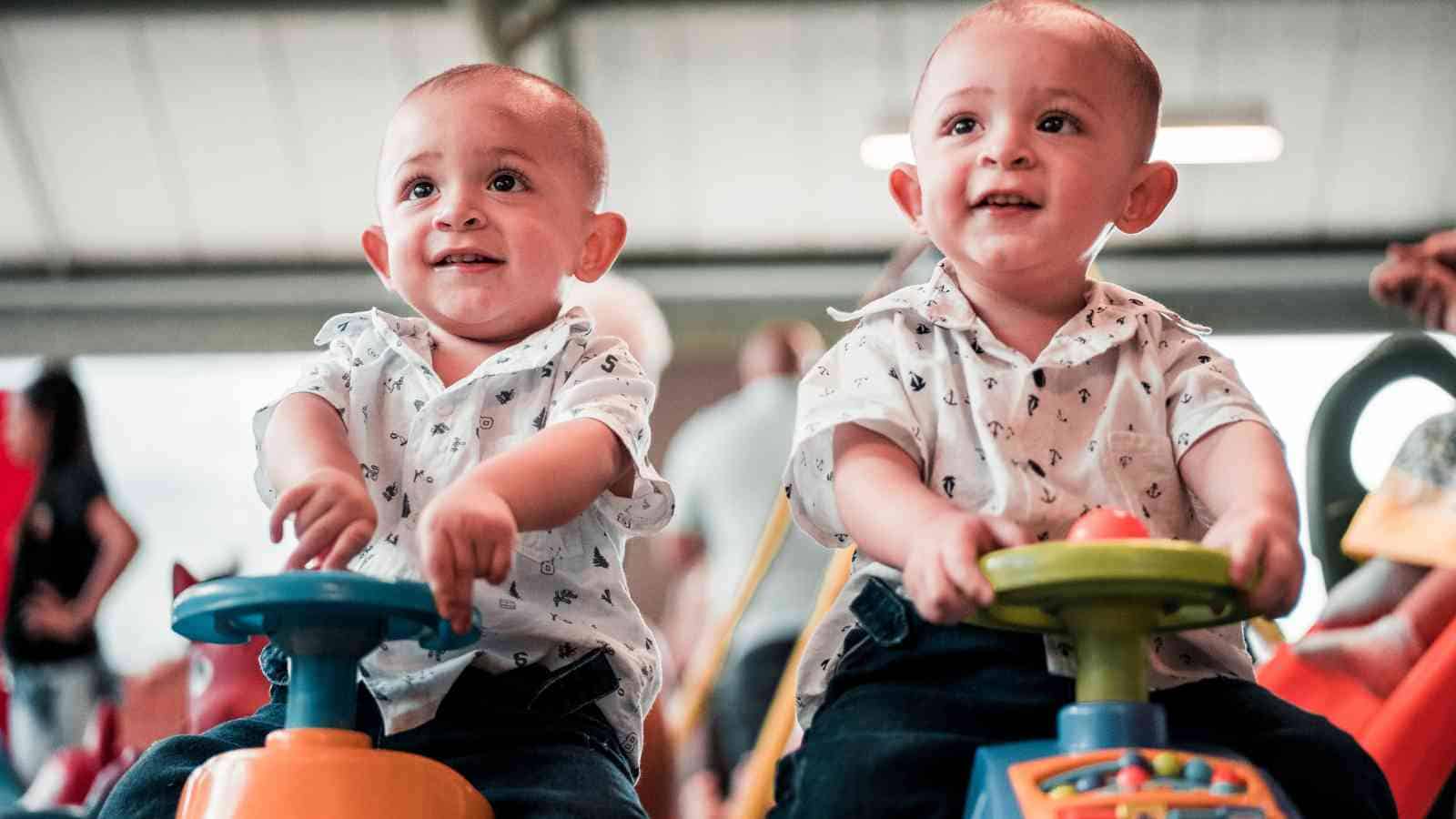 Two young children, dressed in matching white shirts, sit on toy cars in an indoor play area.