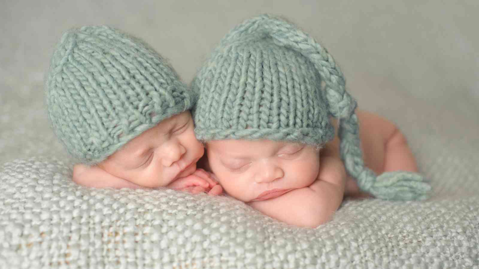 Two sleeping babies wearing knitted hats lie on a soft, textured blanket.