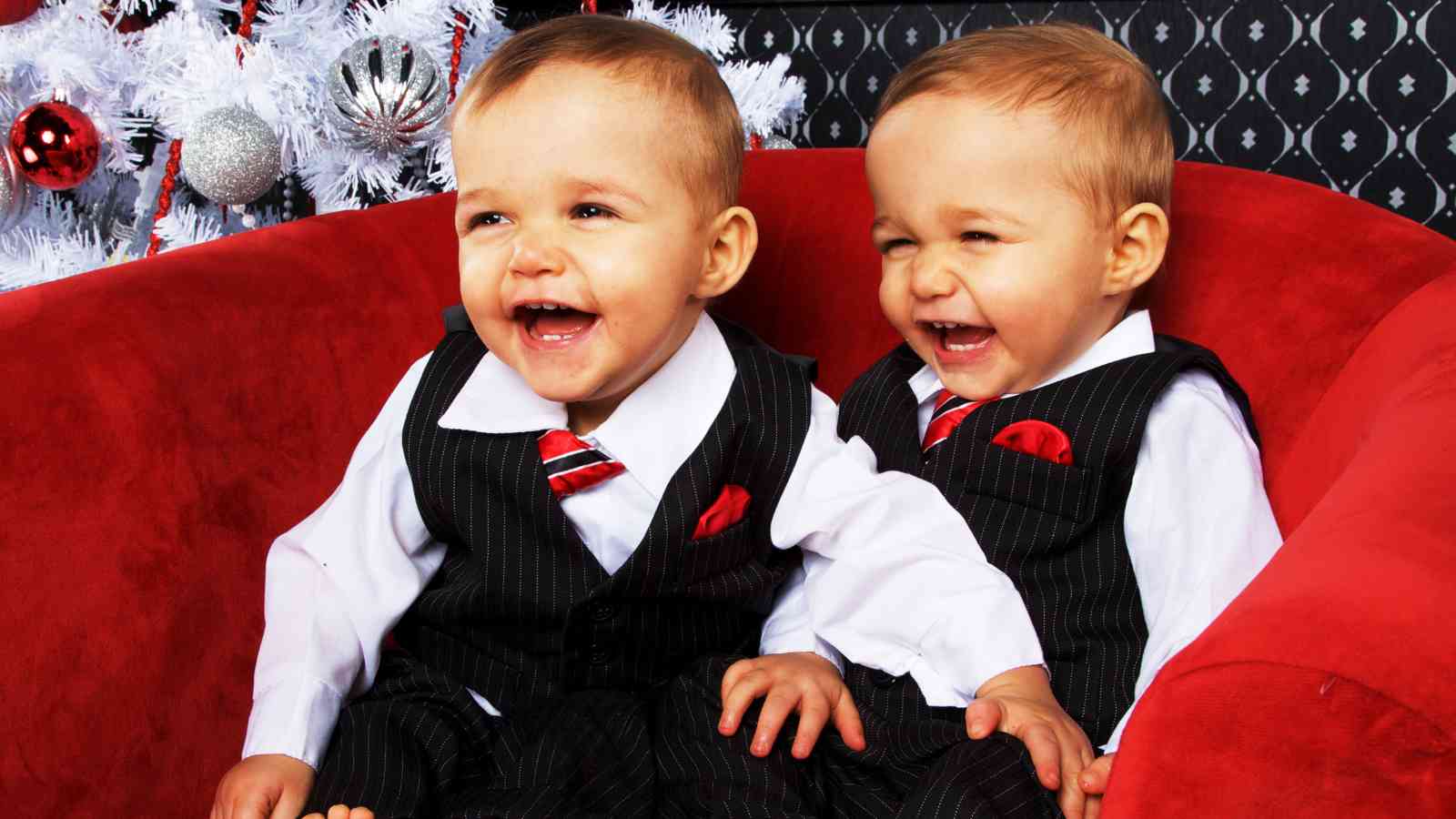 Two young children wearing matching pinstripe suits and red ties sit on a red chair, smiling and laughing in front of a white Christmas tree.