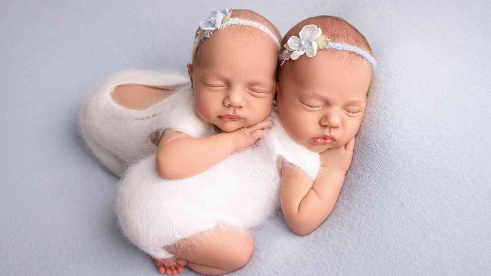 Two newborn baby girls are sleeping soundly, side by side, on a light blue blanket.