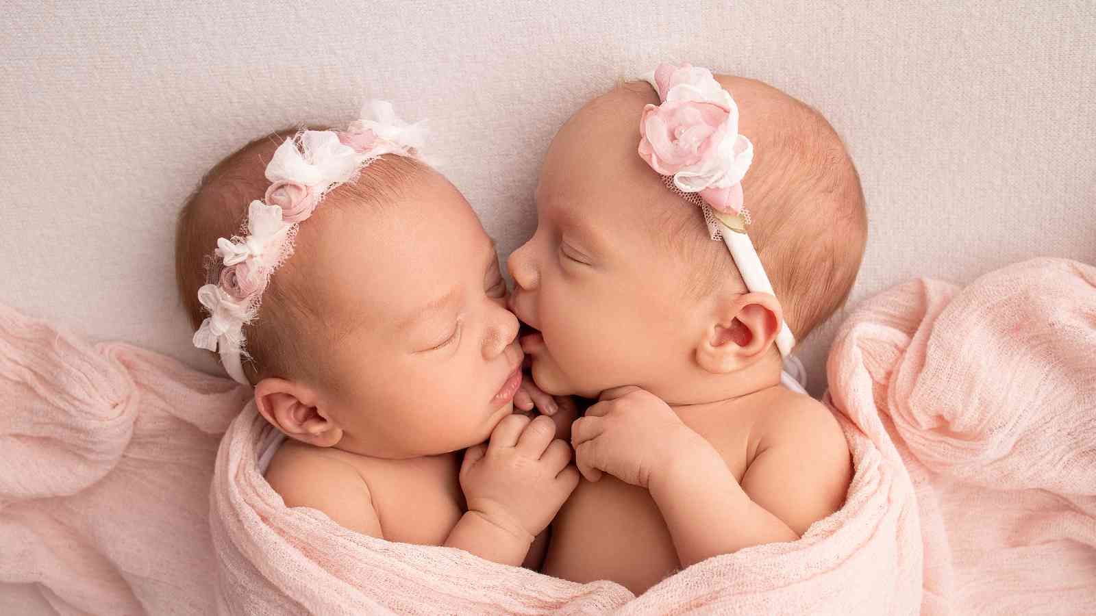 Two baby girls wearing flower headbands are sitting in a pink basket.
