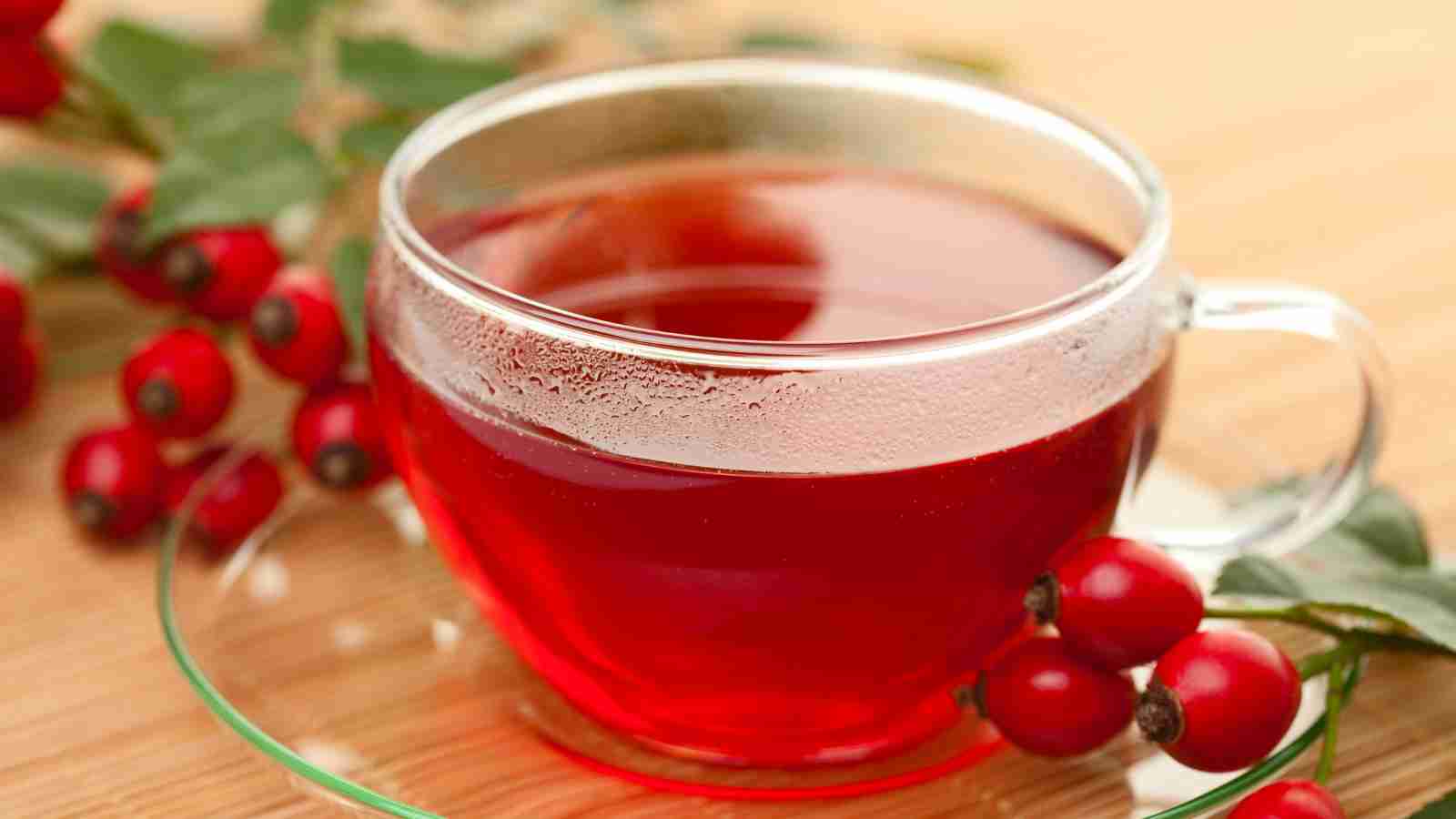 A clear glass cup filled with red rosehip tea is placed on a glass saucer, surrounded by fresh rosehips and green leaves on a wooden surface.