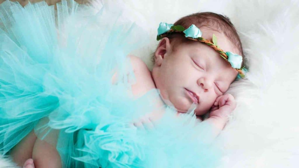 A newborn baby girl is sleeping peacefully on a white fluffy blanket wearing a light blue tutu and a flower crown.
