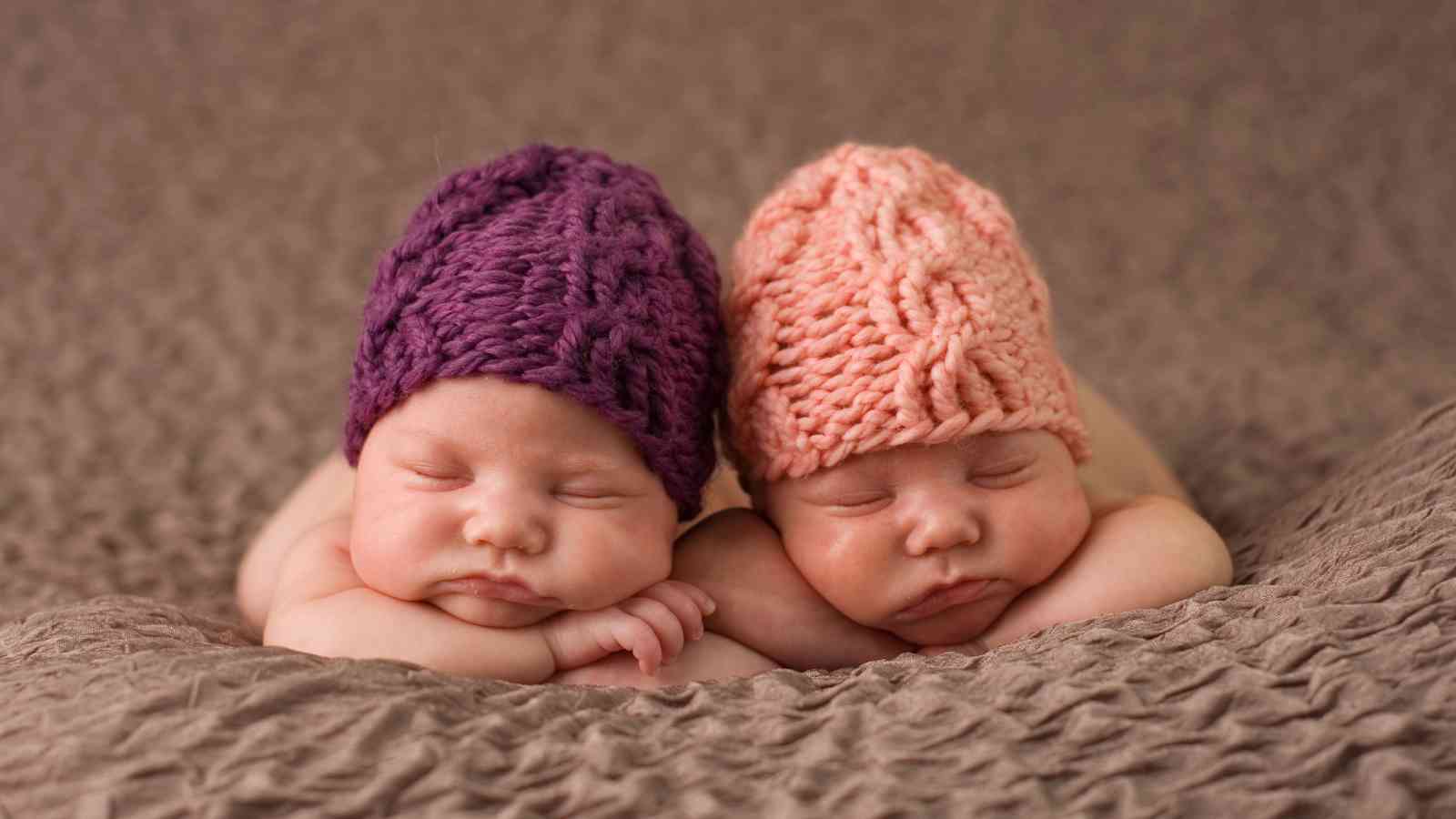 Two newborn baby girls are sleeping soundly, side by side, on a brown blanket. The baby on the left is wearing a purple knitted hat and the baby on the right is wearing a peach knitted hat.