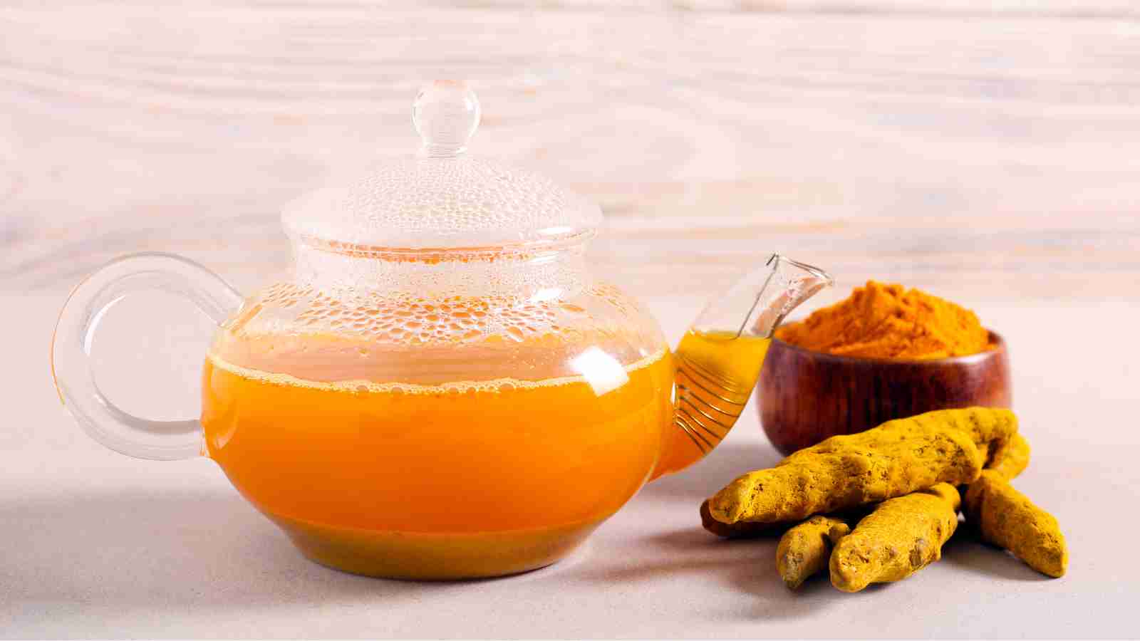 A glass teapot filled with turmeric tea is placed next to a bowl of turmeric powder and several turmeric roots on a light background.