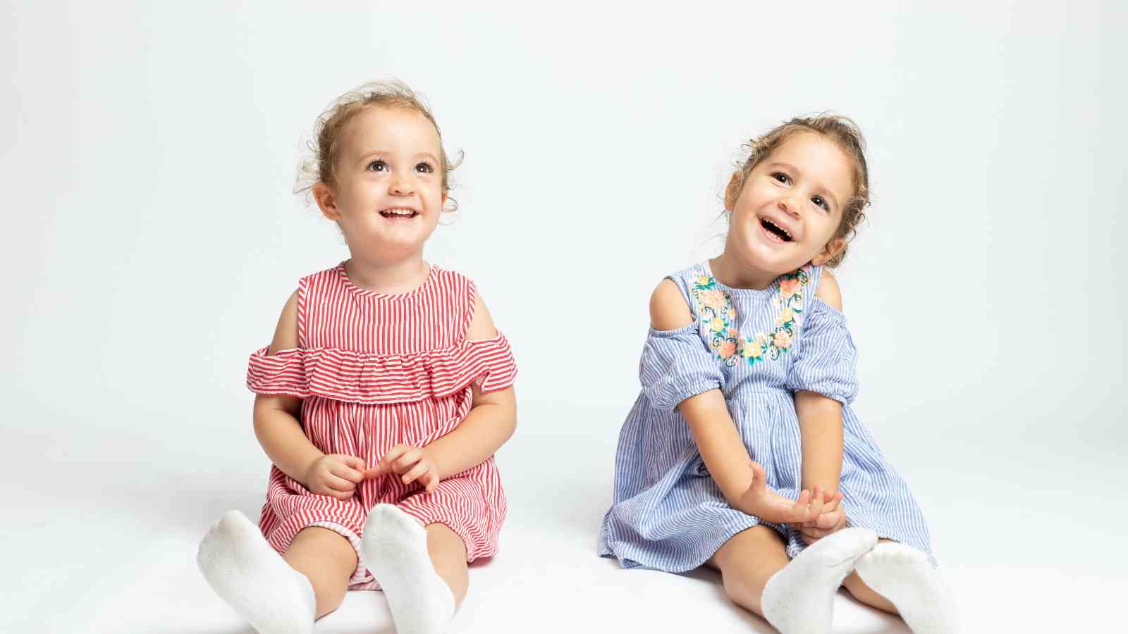 Twin girls sitting on a white surface wearing matching striped dress.