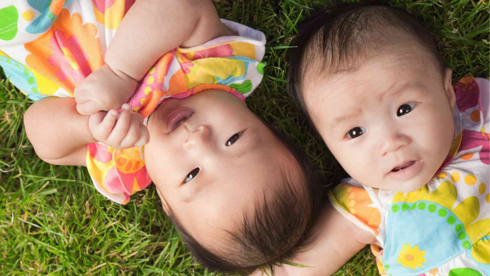 Two baby girls in matching floral dresses are lying on their backs on a patch of green grass.