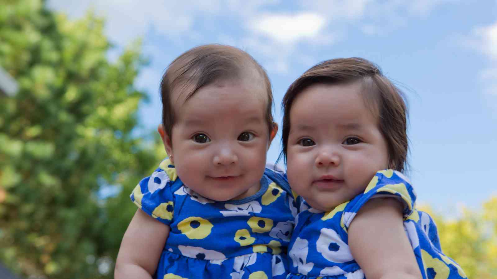 Two baby girls, wearing matching blue dresses, are looking at the camera.