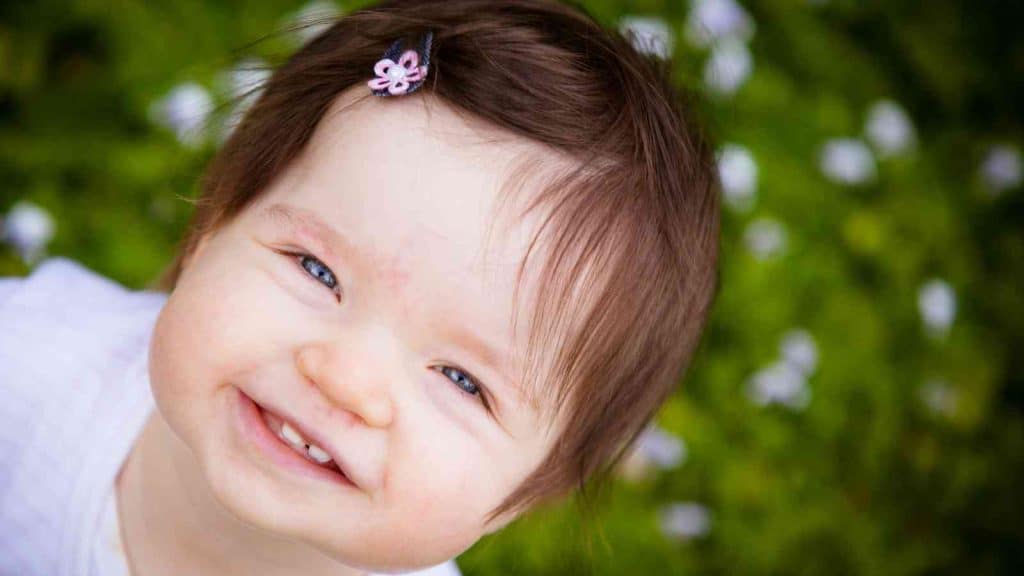 A close-up photo of a baby girl with brown hair and blue eyes, wearing a white shirt and has a flower clip in her hair.