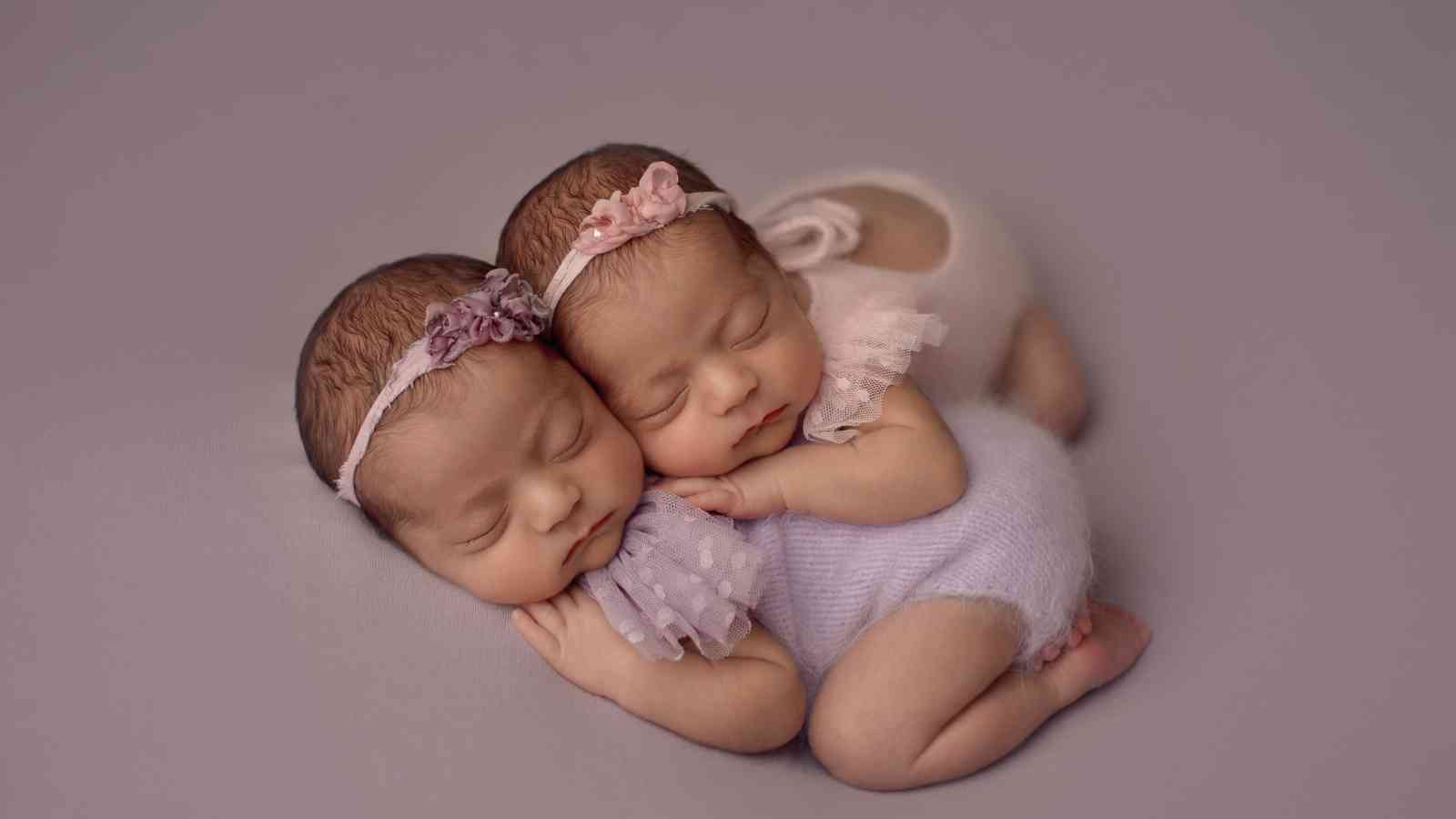 Two newborn babies wearing headbands and matching outfits are sleeping together on a purple background.