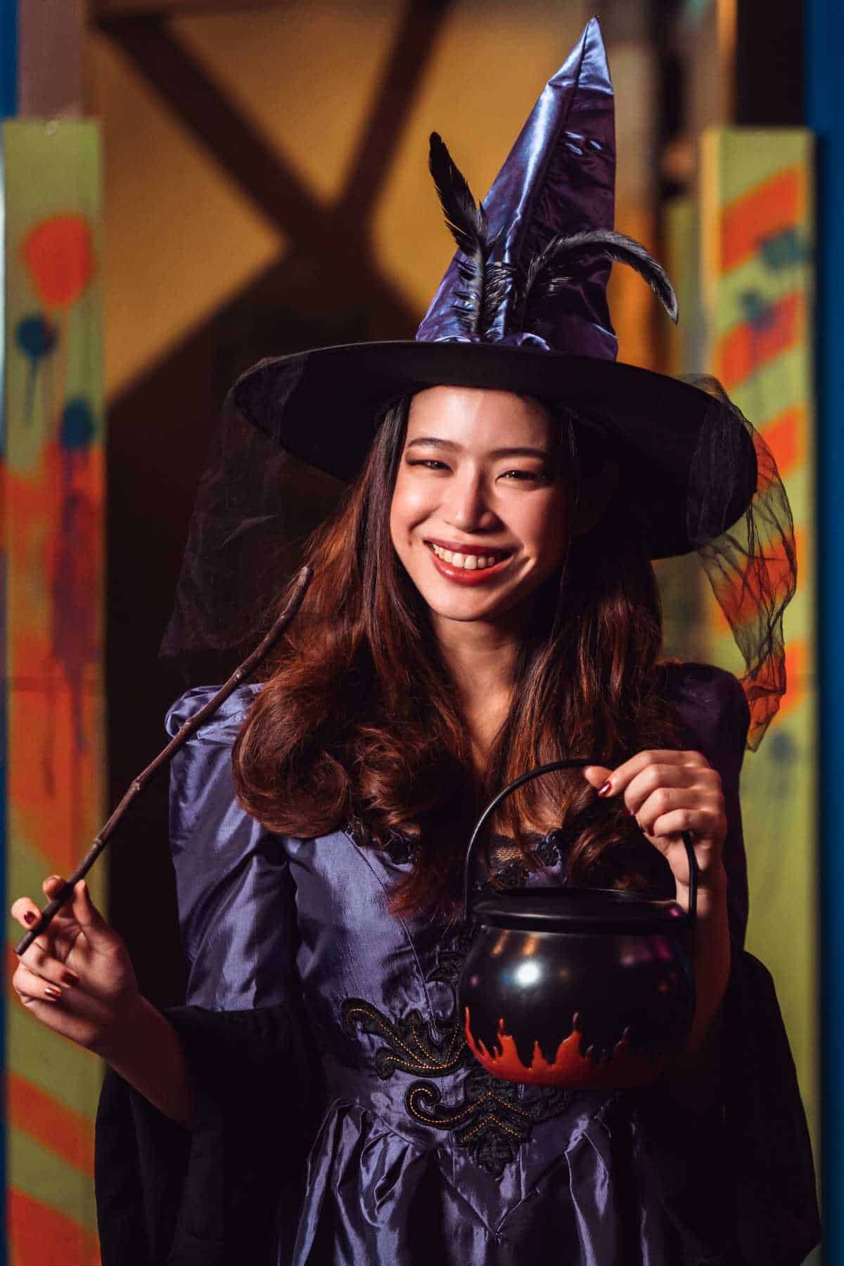 A woman in a witch costume holding a wand and a cauldron-shaped bucket, smiling.