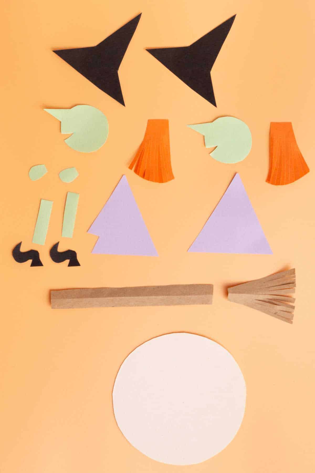 Cut-out paper pieces on a flat surface including a witch's hat, hair, shoes, and a broomstick, arranged on an orange background.