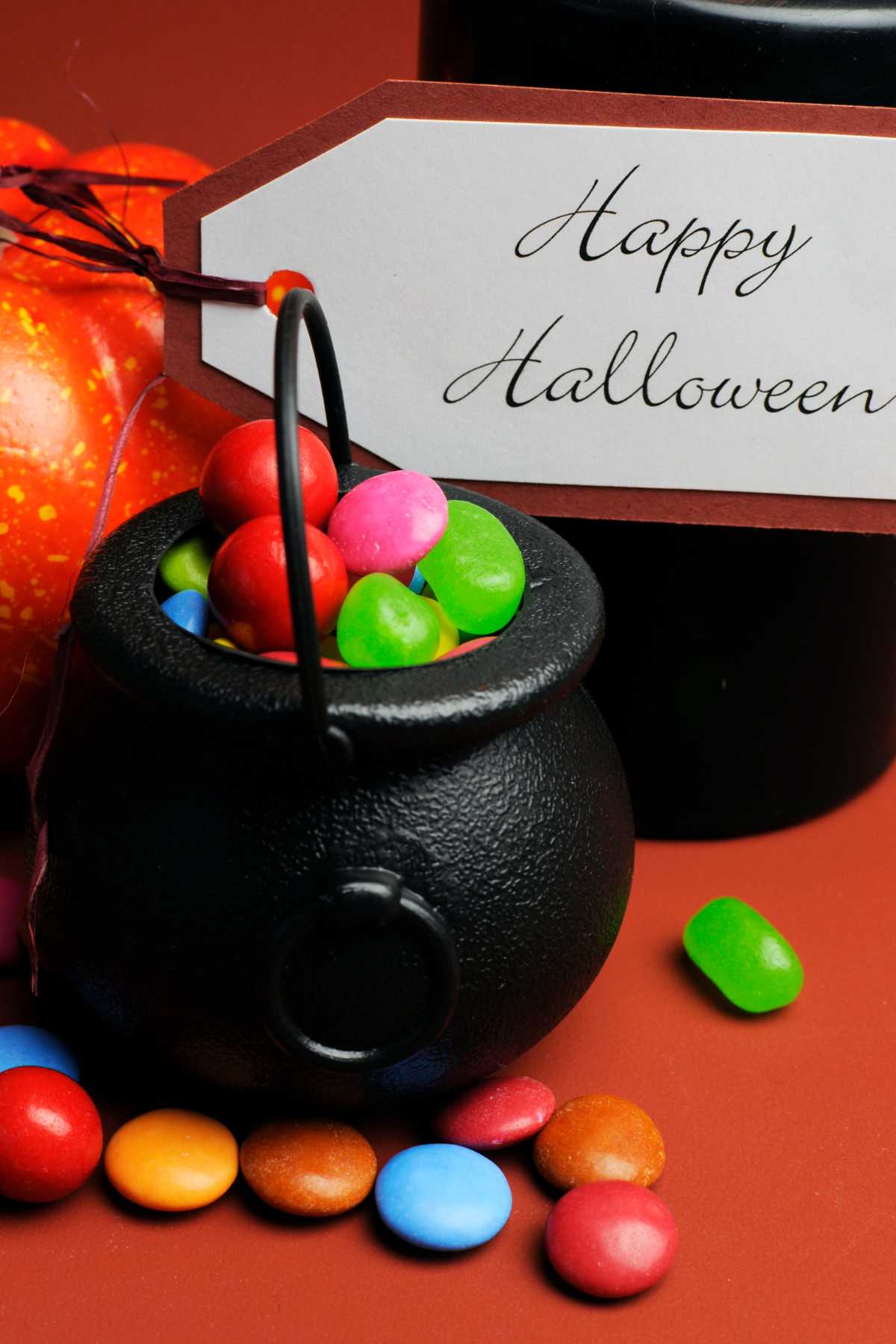 A small black cauldron filled with colorful candies is placed on a surface with more candies scattered around, alongside a "Happy Halloween" tag.