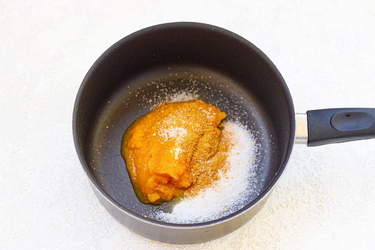 A black saucepan filled with a dollop of orange pumpkin puree, granulated sugar, and pumpkin spice on a light surface.